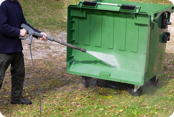 Person pressure washing a green dumpster.
